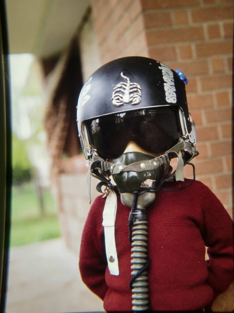 Bernardo bambino, a circa 6 anni, in giardino gioca indossando il casco da volo del padre Fausto e ha la visiera nera abbassata sugli occhi e la maschera dell'ossigeno chiusa sul viso