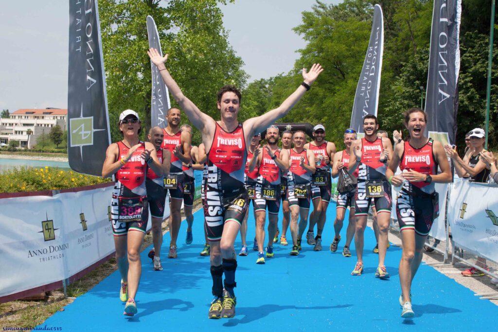 Bernardo con le braccia alzate al traguardo della gara di triathlon alle Bandie, la squadra lo accompagna. si vedono i tutori in carbonio toe off con cui riesce a correre