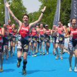 Bernardo felice e sorridente con le braccia alzate al traguardo della gara di triathlon alle Bandie, la squadra lo accompagna. si vedono i tutori in carbonio toe off con cui riesce a correre