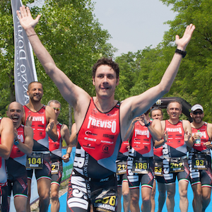 Bernardo con le braccia alzate al traguardo della gara di triathlon alle Bandie, la squadra lo accompagna. si vedono i tutori in carbonio toe off con cui riesce a correre