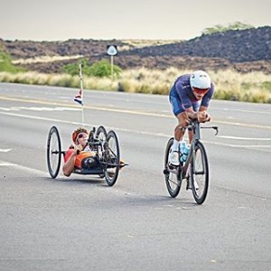 Nella foto si vedono due atleti che stanno svolgendo la finale del campionato del mondo di Ironman a Cona, nelle Hawai. Sono un atleta normo dotato e un atleta disabile, impegnati nella frazione di bicicletta
